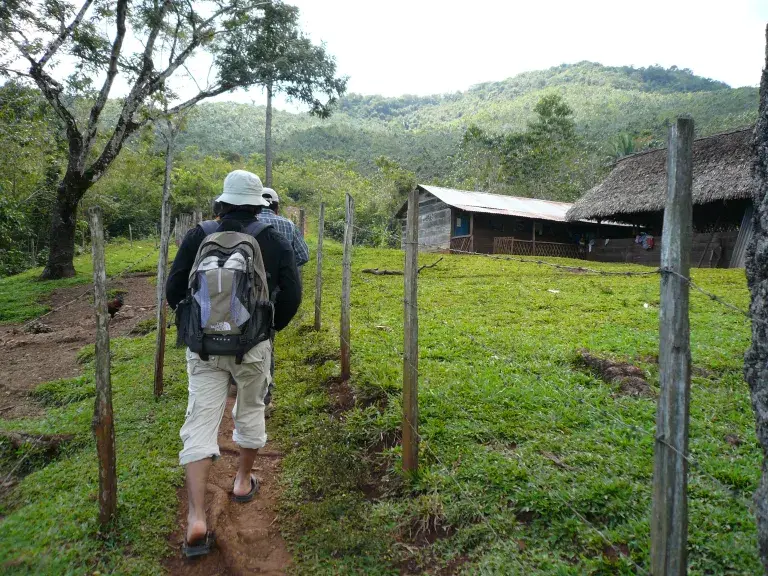 Hombre indígena que camina por un camino de tierra al lado de un área agrícola cercada en el área agrícola