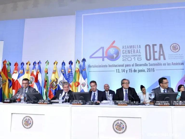 Image shows 10 people at a conference table, sitting in front of multiple Nations flags behind them. There is a logo projected on the wall that reads: 46th Assembly General Dominican Republic 2016 Institutional Strengthening for Sustainable Development in the Americas June 13,14, and 15, 2026.